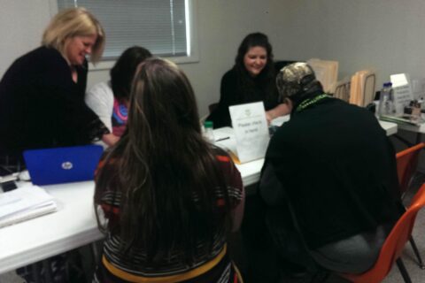 Group of people seated at a table during a medical cannabis outreach event, with a “Please check in here!” sign and registration materials visible.
